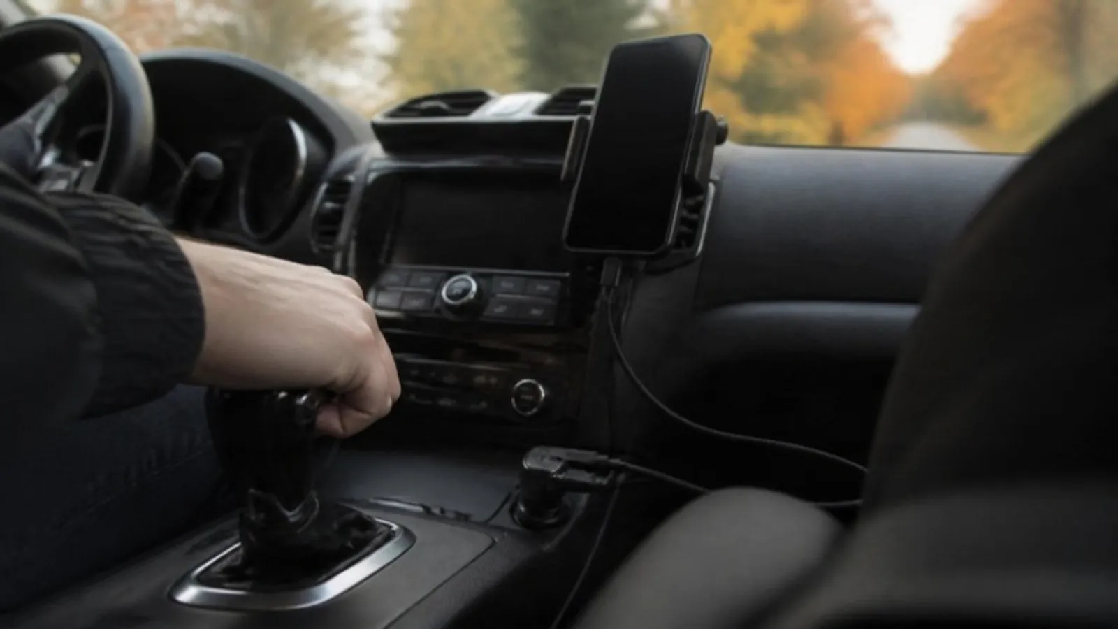 A smartphone charging from a USB-C cable plugged into a car's 12V socket charger on the center console during a daytime drive