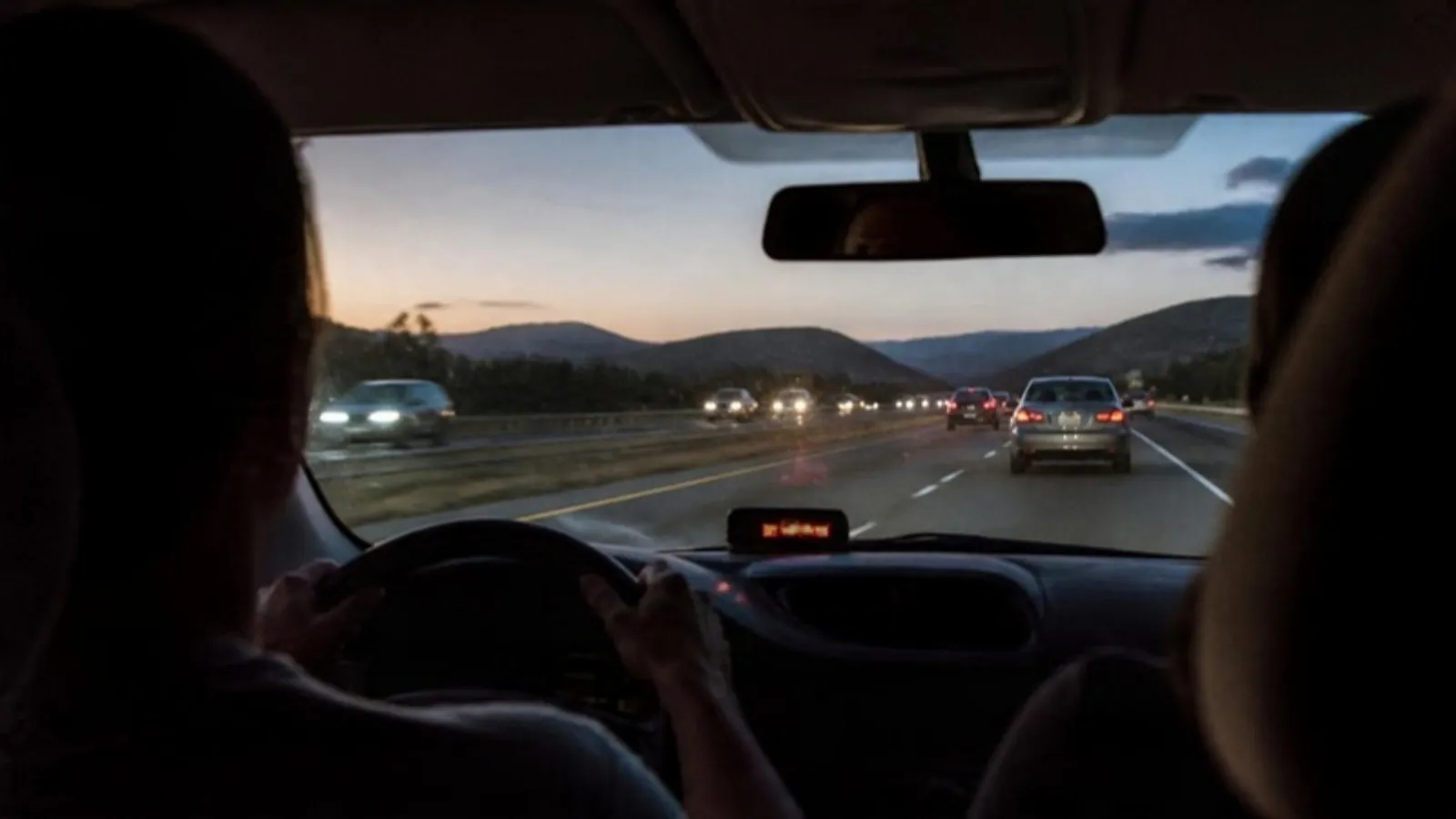 A small speed display glowing on a car dashboard below the windshield while a driver travels on a highway at dusk