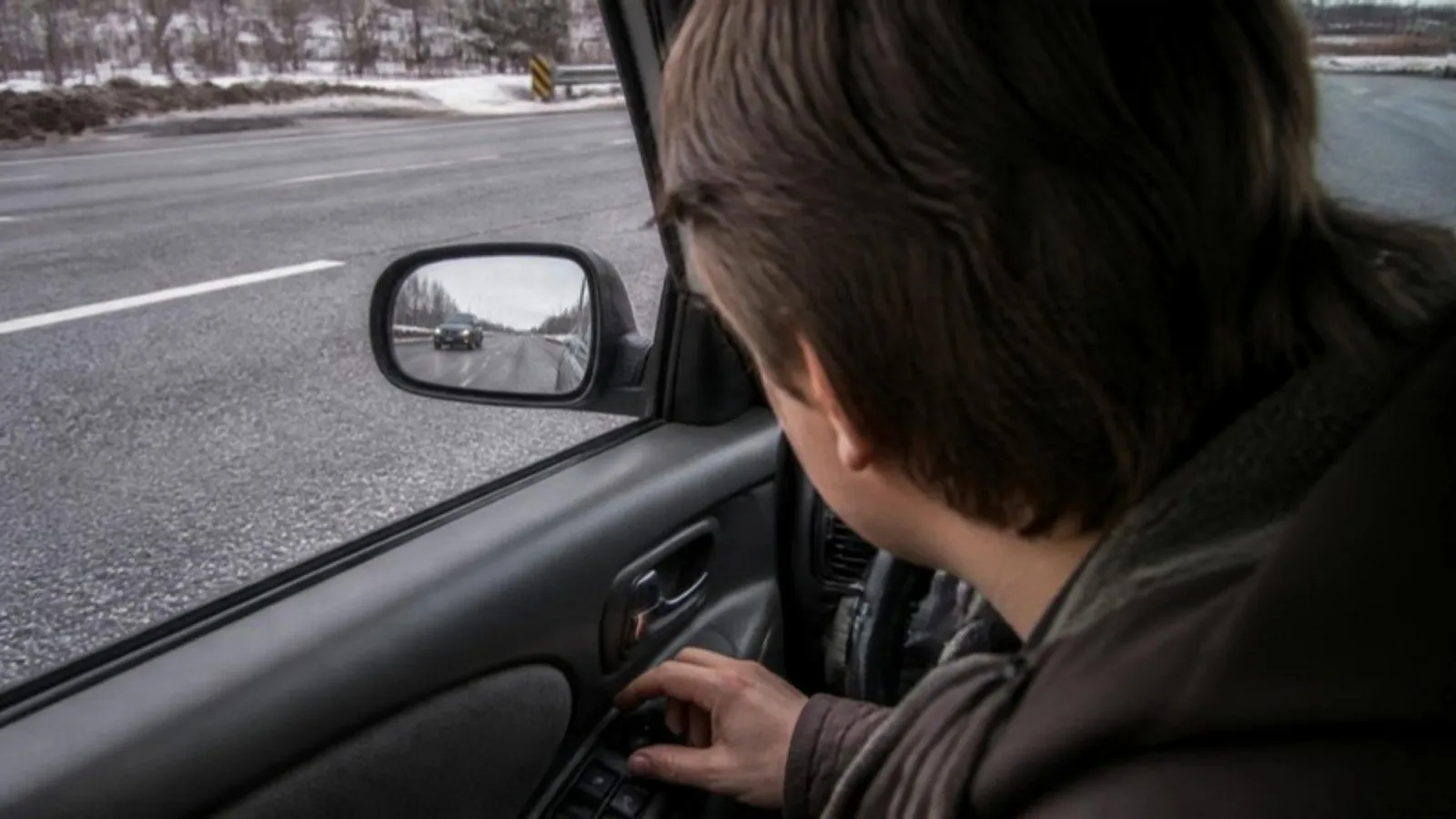 Driver leaning toward the window to adjust a side mirror while a car passes in the next lane on a gray winter day
