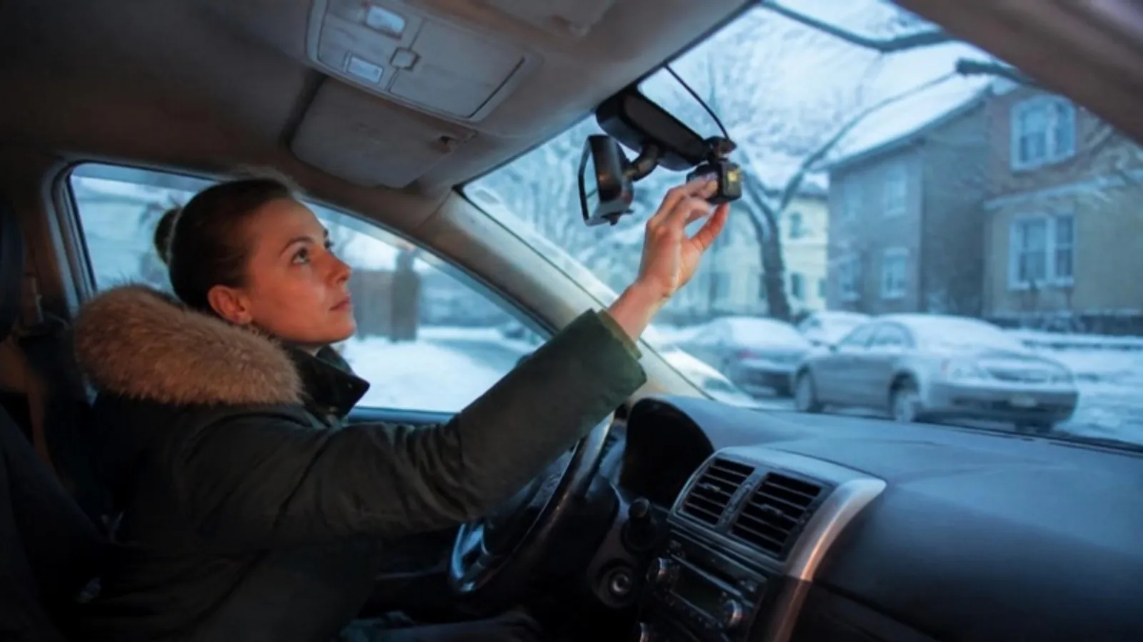 Driver adjusting a small dash cam behind the rearview mirror of a car parked on a snowy street at dusk