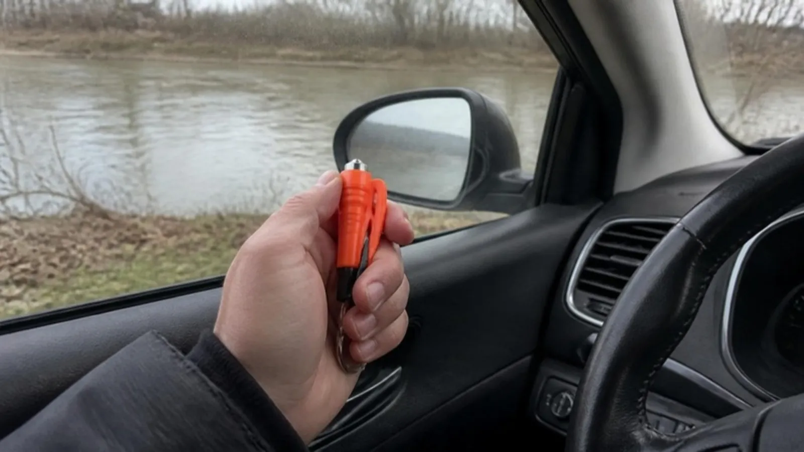 A driver's hand holding a small window breaker tool next to the driver's side window of a car parked by a river